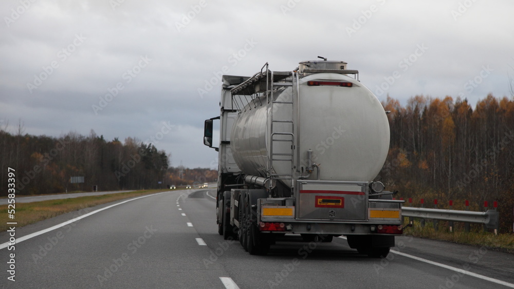 Food liquids transportation, semi-truck with barrel move on suburban empty asphalted road at autumn evening on gray sky background, liquid goods transportation