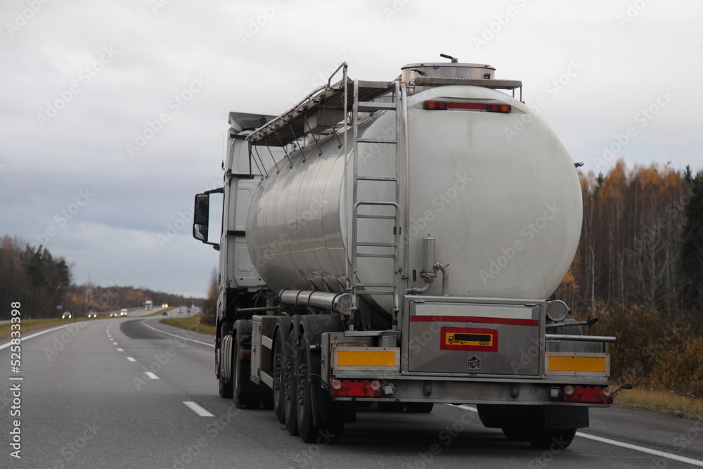 Big semi truck with barrel for liquid food goods drive on suburban highway road at autumn evening in perspective, back side view