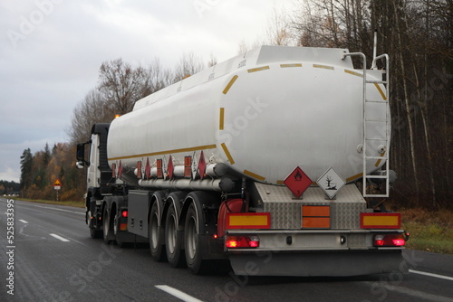 European tree-axle semi truck fuel tanker move on suburban highway road at autumn evening in perspective, back side view, gasoline fuel ADR dangerous cargo transportation logistics in Europe