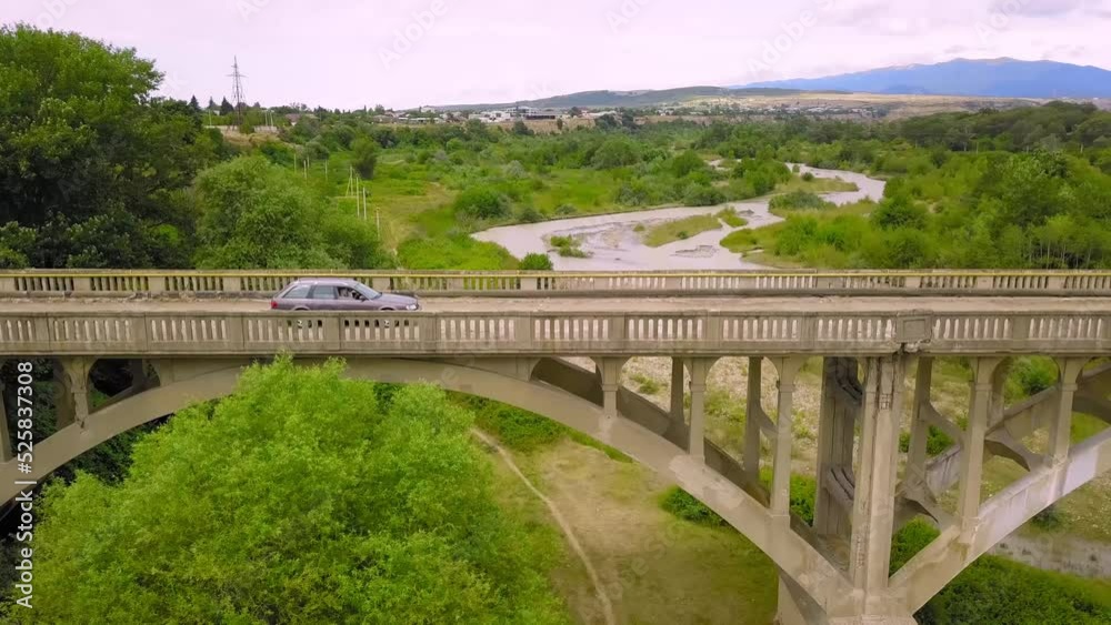 Blue car crosses river on old abandoned bridge against backdrop of mountains. Car rides on old arch bridge over mountain river on summer day, side view from drone. Georgia. Road trip. Blue car crosses river on old abandoned bridge against backdrop of mountains. Car rides on old arch bridge over mountain river on summer day, side view from drone. Georgia. Road trip.