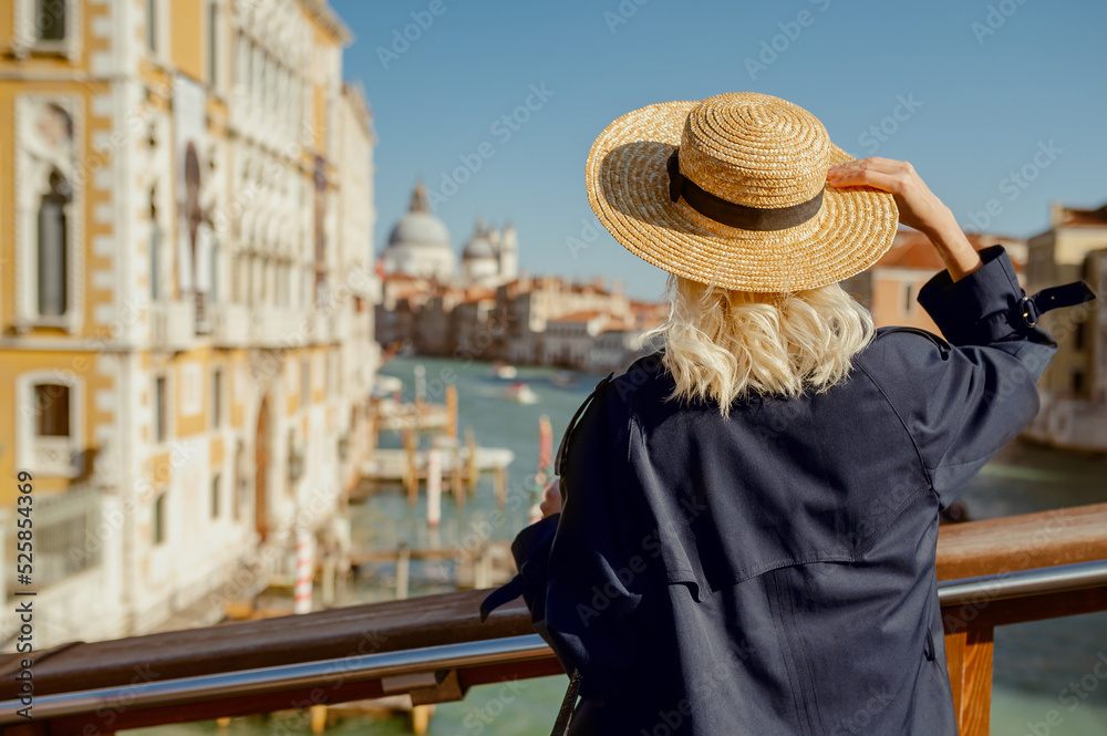 Rear, back view of traveler woman wearing straw hat, autumn trench coat, posing on Academia bridge with great view on Grand canal in Venice. Travel, vacation, lifestyle conception. Copy, empty space