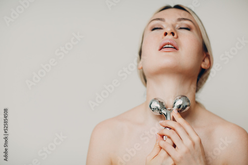 Beautiful young adult woman with massage face roller on beige background