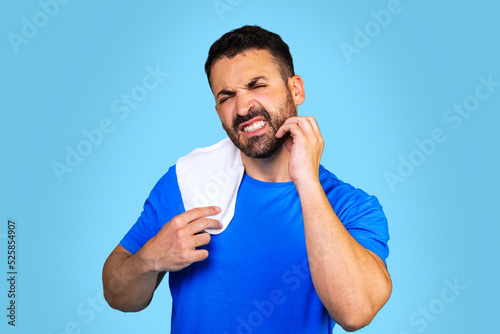  A bearded caucasian man in a blue t-shirt with a towel scratches his beard on a blue background. Irritating itching.