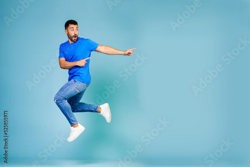 Full length portrait of cheerful young man jumping in blue t-shirt pointing finger to the side celebrating success against blue background