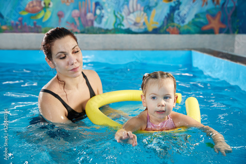 Preschool girllearning to swim in pool with foam noodle with young trainer photo