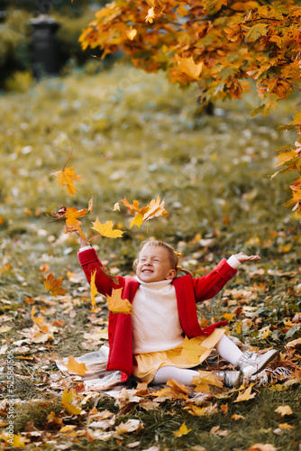 Portrait of a happy smiling little girl child in a bright red coat catching autumn leaves having fun and laughing sitting on a plaid blanket on the foliage in an autumn park outdoors in fall. 