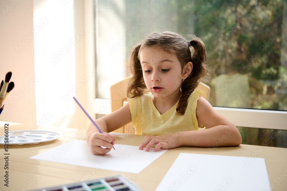 Adorable little preschooler girl in yellow t-shirt and two ponytails, sitting at a table and drawing picture with a pencil on white blank paper sheet, enjoying art class after school. Kids development Adorable little preschooler girl in yellow t-shirt and two ponytails, sitting at a table and drawing picture with a pencil on white blank paper sheet, enjoying art class after school. Kids development