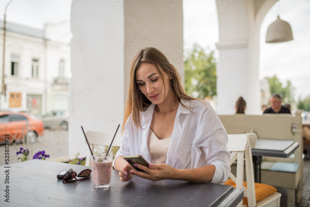 A young attractive girl is holding a smartphone in her hands, checking social communication in a cafe on the street. A woman in casual clothes enjoys using technology during a refreshing cocktail