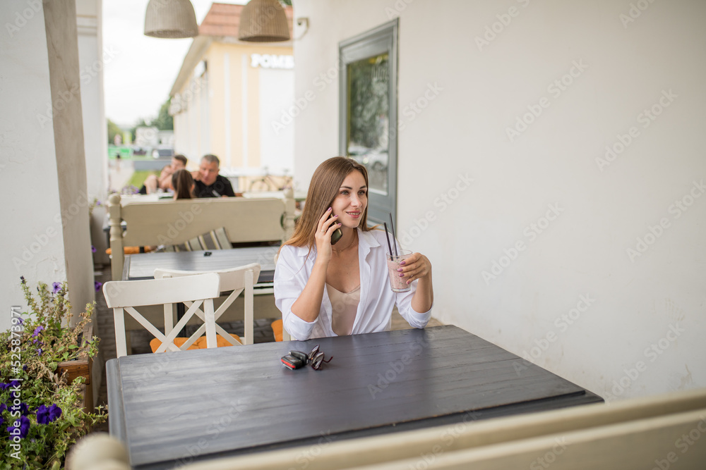 A young attractive girl is holding a smartphone in her hands, checking social communication in a cafe on the street. A woman in casual clothes enjoys using technology during a refreshing cocktail A young attractive girl is holding a smartphone in her hands, checking social communication in a cafe on the street. A woman in casual clothes enjoys using technology during a refreshing cocktail