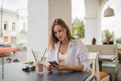 A young attractive girl is holding a smartphone in her hands, checking social communication in a cafe on the street. A woman in casual clothes enjoys using technology during a refreshing cocktail