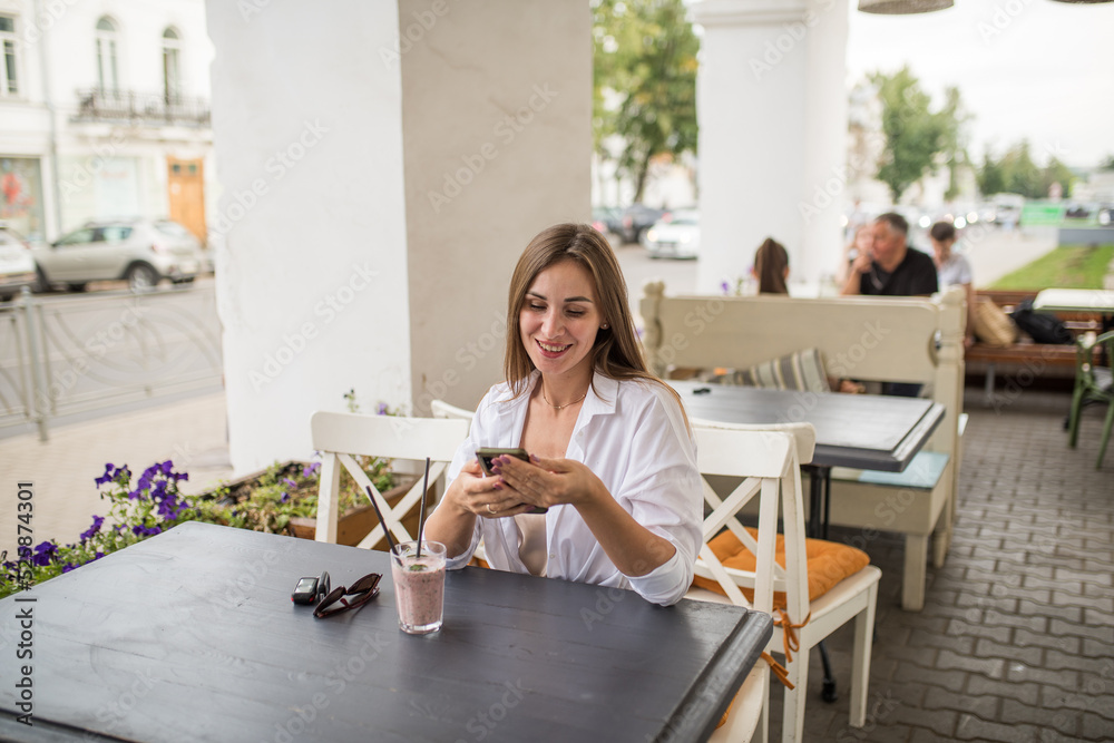 A young attractive girl is holding a smartphone in her hands, checking social communication in a cafe on the street. A woman in casual clothes enjoys using technology during a refreshing cocktail A young attractive girl is holding a smartphone in her hands, checking social communication in a cafe on the street. A woman in casual clothes enjoys using technology during a refreshing cocktail
