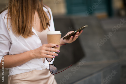 A young woman with a smartphone and coffee in a cup in an urban area of the city, a girl communicates on a mobile phone on the street in summer