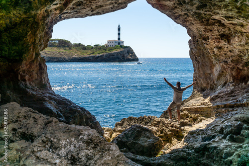 Unrecognizable man with raised hands inside a cave overlooking the sea and a red and white lighthouse with a ship sailing