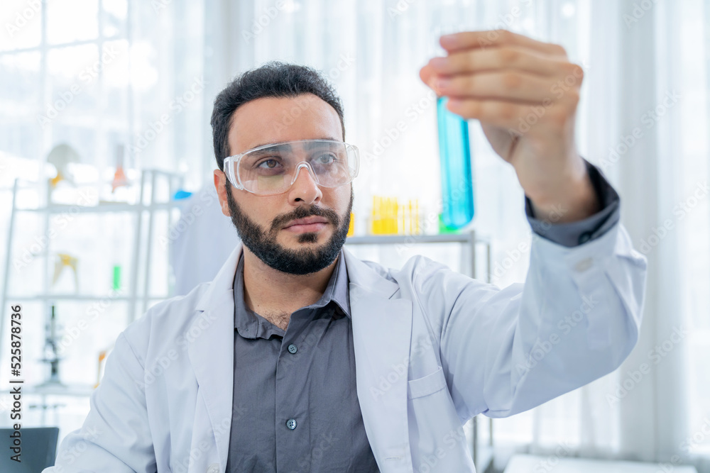 Scientist in lab coat and safety glasses holding test tube with blue solution at modern laboratory. Look at tube and analysis. Research development, bio chemistry, medical and pharmaceutical Scientist in lab coat and safety glasses holding test tube with blue solution at modern laboratory. Look at tube and analysis. Research development, bio chemistry, medical and pharmaceutical