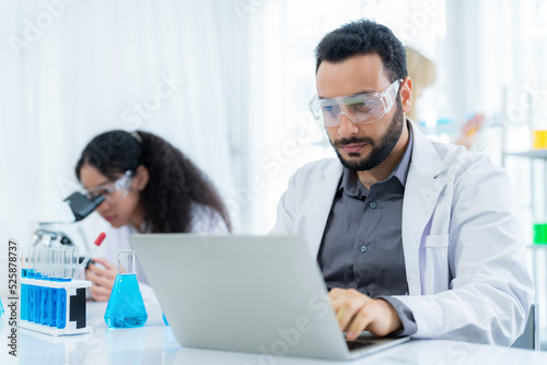 Scientist in lab coat and safety glasses holding flask with blue solution at modern laboratory. Take note on the laptop for analysis. Research development, bio chemistry, medical, pharmaceutical
