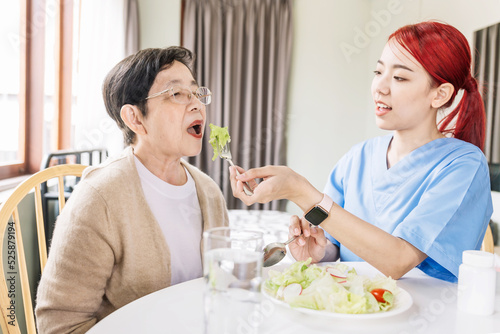 Asian woman caregiver wearing medical scrub takes care of senior Asian woman by feeding vegetarian salad to eat at home. caregivers visit home Home health care concept and nursing home.