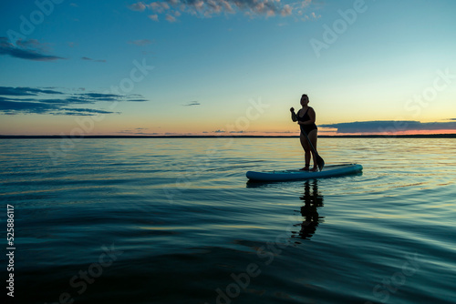 a woman in a closed swimsuit with a mohawk standing on a SUP board with an oar floats on the water at dusk.