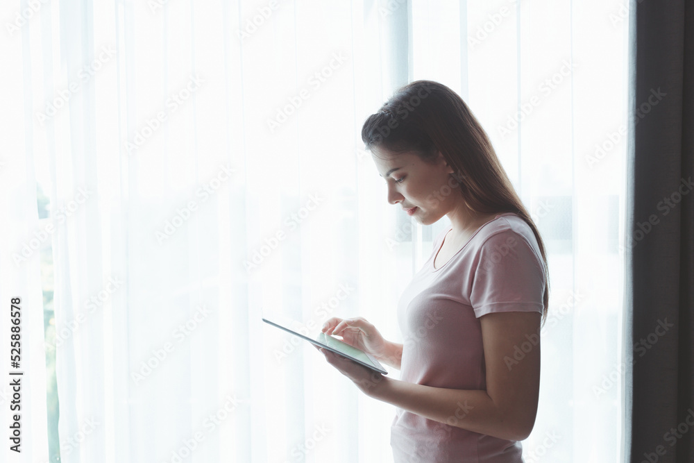 Asian businessman works from home using a tablet device, browsing the Internet, playing social media, Attractive businesswoman using digital tablet while standing in front of a window in home Asian businessman works from home using a tablet device, browsing the Internet, playing social media, Attractive businesswoman using digital tablet while standing in front of a window in home