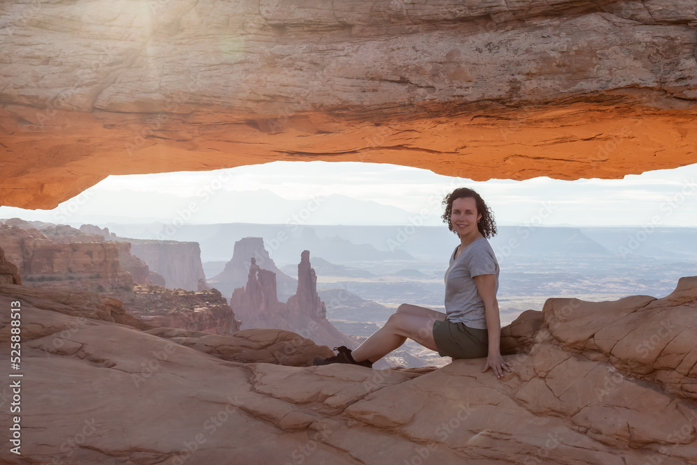 Adventurous Woman at a Scenic American Landscape and Red Rock Mountains in Desert Canyon. Spring Season. Sunrise Sky. Mesa Arch in Canyonlands National Park. Utah, United States. Adventure Travel