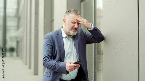 Mature business man with dizziness, vertigo a severe headache standing on a city street leaning against a wall. outside. Senior male with high blood pressure or stroke holding head massaging. Outdoor photo