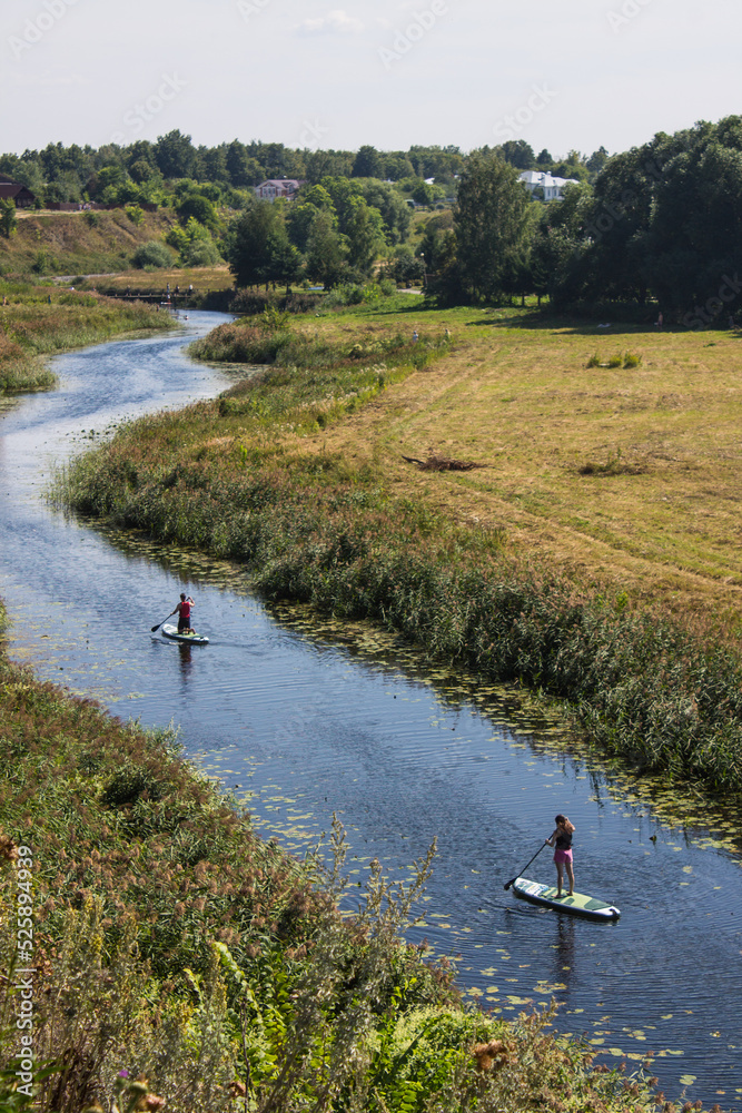 Tourists are rafting on the winding Kamenka river in Suzdal Russia among meadows with grass on a sunny summer bright day and a space for copying Tourists are rafting on the winding Kamenka river in Suzdal Russia among meadows with grass on a sunny summer bright day and a space for copying