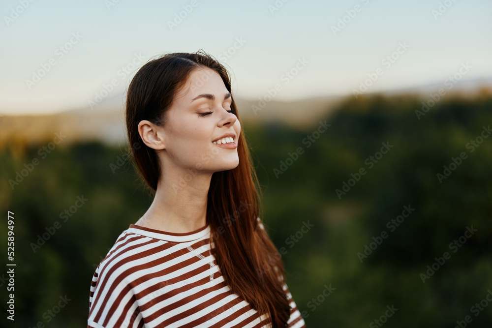 Woman traveler in nature in a striped t-shirt smiling and looking at the sunset