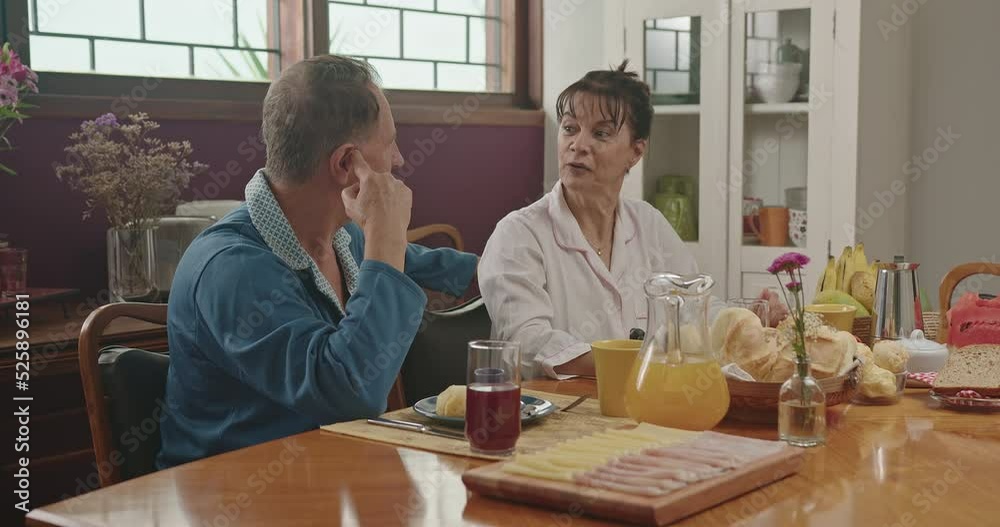 A middle aged couple chatting during breakfast. They are sitting at the lavish kitchen table, smiling and wearing pajamas. Married couple in conversing in the morning breakfast table A middle aged couple chatting during breakfast. They are sitting at the lavish kitchen table, smiling and wearing pajamas. Married couple in conversing in the morning breakfast table