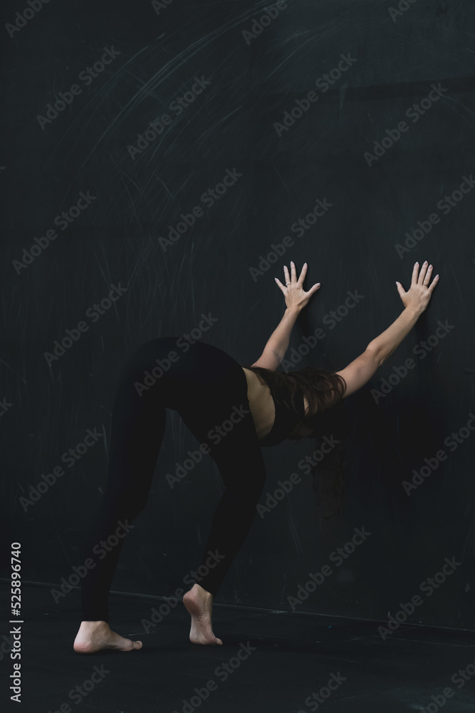 Young woman meditates, doing yoga poses and asana. Fitness girl enjoying yoga indoors. A yoga gymnast girl on a dark background. Black on Black. Young woman meditates, doing yoga poses and asana. Fitness girl enjoying yoga indoors. A yoga gymnast girl on a dark background. Black on Black.