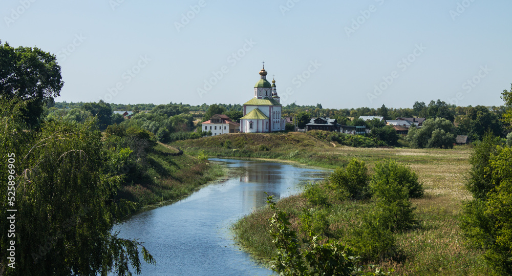 The white stone Church of Elijah the Prophet on the bank of the Kamenka river in Suzdal Russia on a summer day and old houses among lush green foliage