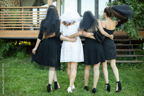 Girls in dresses stand with their backs on green grass. Maidens with bride in white dress. Black dresses on girlfriends.