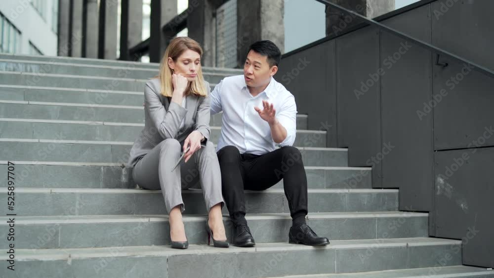 Asian male employee supports upset depressed female co-worker colleague. Sitting on the steps stairs outside an office building. young man comforting sad woman at work outdoors Asian male employee supports upset depressed female co-worker colleague. Sitting on the steps stairs outside an office building. young man comforting sad woman at work outdoors