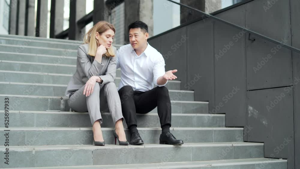 Asian male employee supports upset depressed female co-worker colleague. Sitting on the steps stairs outside an office building. young man comforting sad woman at work outdoors Asian male employee supports upset depressed female co-worker colleague. Sitting on the steps stairs outside an office building. young man comforting sad woman at work outdoors