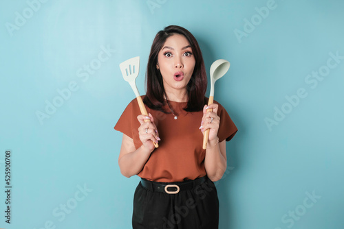 Slika na platnu Excited Asian woman holding cooking ware and smiling, isolated by blue backgroun