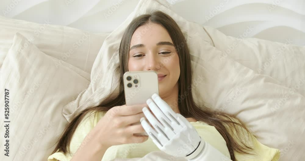 Girl with bionic arm using phone, reading social media and chatting in bed, woman with disability learning or working online remotely from home Girl with bionic arm using phone, reading social media and chatting in bed, woman with disability learning or working online remotely from home