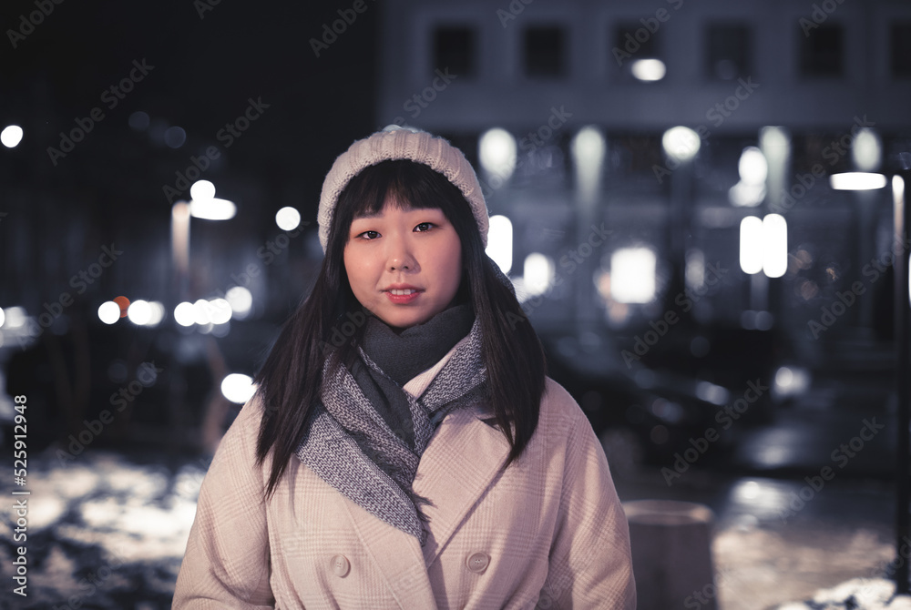 a young Korean woman in a warm hat on a city street. A happy carefree girl in a beige fashionable coat and standing on the street on a winter evening in the city center at dusk on a walk.