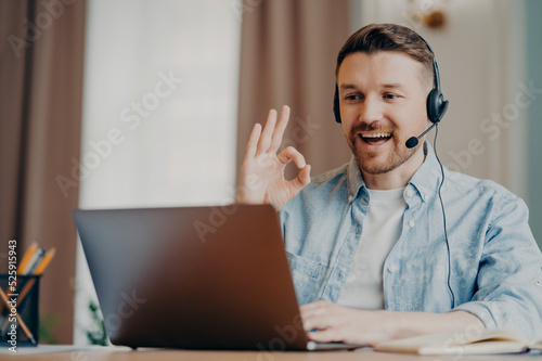Smiling stylish man in headset showing ok gesture while having video call