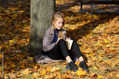 a beautiful girl in a plaid coat stands leaning on a tree trunk in an autumn park. holds a bouquet of yellow maple leaves in her hands and looks intently to the side