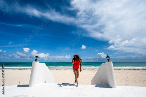 Woman on vacation in Mexico wearing a red bathing suit and sun hat holding a glass of champagne walking on beautiful white sand beach with turquoise blue green ocean and blue sky