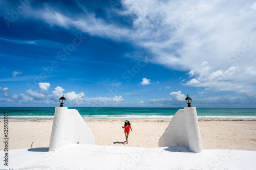 Woman on vacation in Mexico wearing a red bathing suit and sun hat walking on beautiful white sand beach with turquoise blue green ocean and blue sky