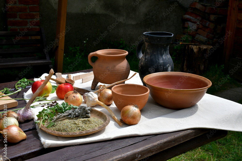 A view of a wooden table full of medieval style dishes and condiments including carrots, ground pepper, basil, thyme, rice, flour, and many other foodstuffs seen on a sunny summer day in a garden
