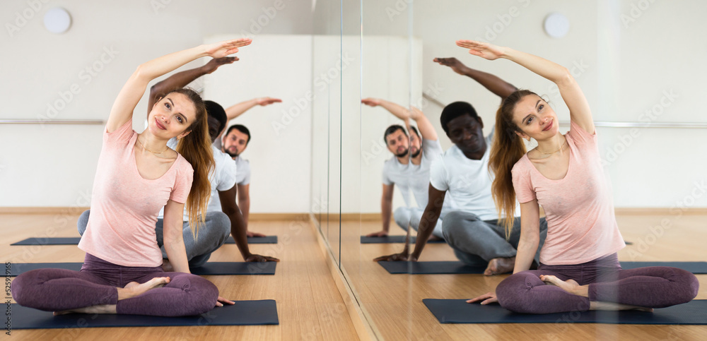 custom made wallpaper toronto digitalSmiling young woman practicing yoga during group class in fitness studio, doing side bend stretching in Padmasana pose. People sitting on mats near mirror with reflection