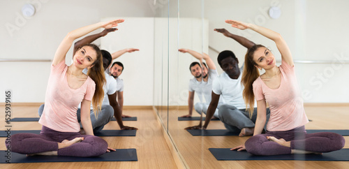 Wallpaper Mural Smiling young woman practicing yoga during group class in fitness studio, doing side bend stretching in Padmasana pose. People sitting on mats near mirror with reflection Torontodigital.ca