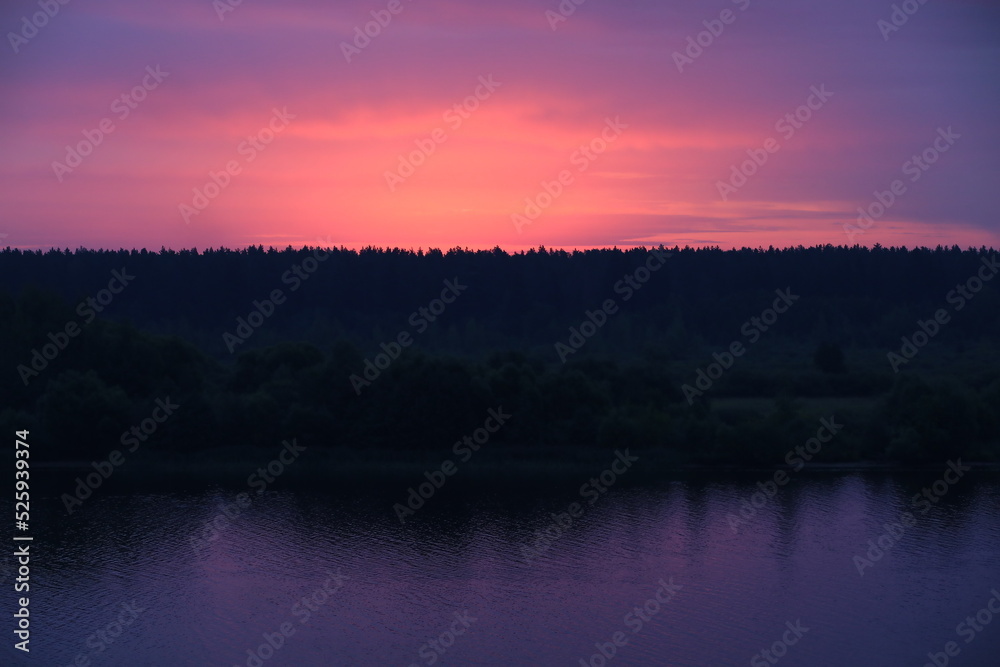 A colorful sunset landscape with a purple sky above the black silhouette of a forest on the riverbank reflected in the lilac water
