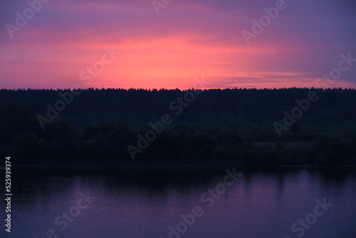 A colorful sunset landscape with a purple sky above the black silhouette of a forest on the riverbank reflected in the lilac water