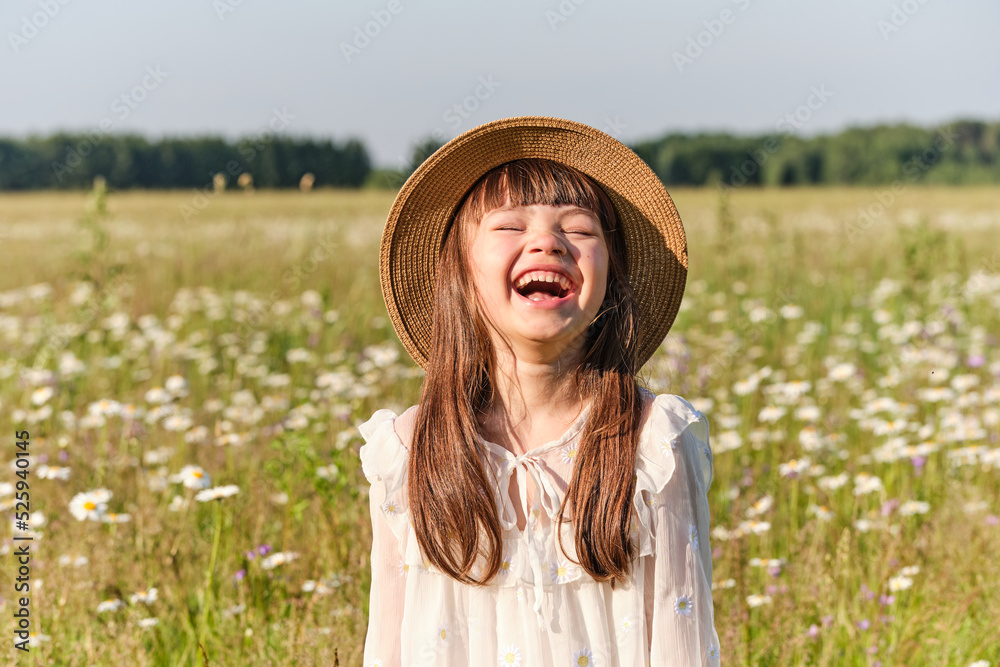 Portrait of a little smiling girl in a white dress and a straw hat on a chamomile field on a bright sunny summer day