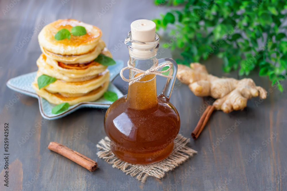 Homemade spicy ginger syrup with honey and cinnamon in glass cruet with plate of fluffy pancakes on background. Homemade spicy ginger syrup with honey and cinnamon in glass cruet with plate of fluffy pancakes on background.