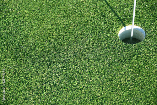 Ein kräftig grüner Golfrasen als Hintergrund mit einem Loch als Ziel. Der Rasen ist kurzgeschnitten und das Loch mit einer Fahne versehen. Die Sportart ist sehr gebliebt. 