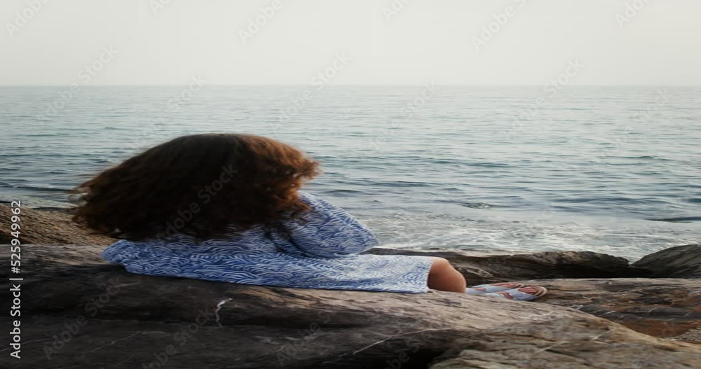 A woman straightens her wavy dark hair, sitting on the rocks on the seashore on a sunny day and looking into distance. Vertical video from back, no face, unrecognizable person. 4k video, red komodo A woman straightens her wavy dark hair, sitting on the rocks on the seashore on a sunny day and looking into distance. Vertical video from back, no face, unrecognizable person. 4k video, red komodo