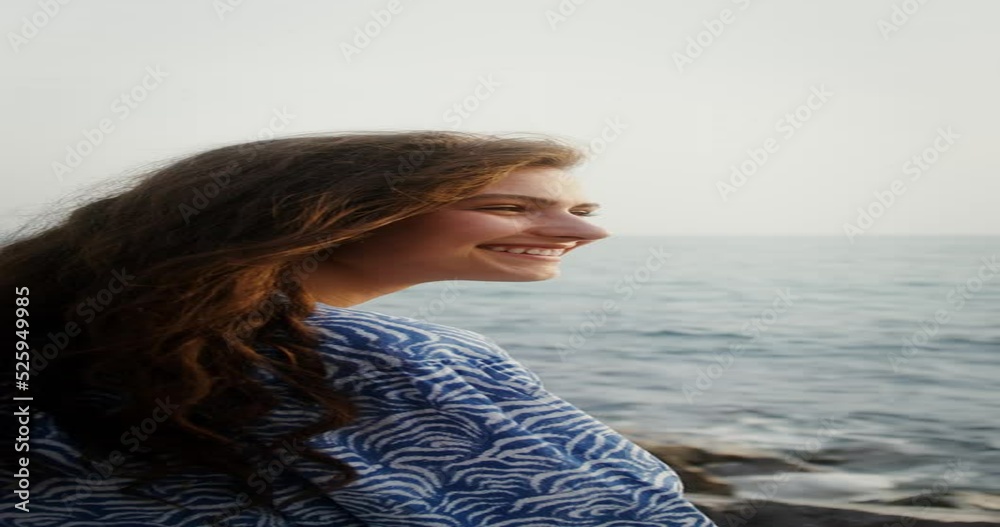 A young beautiful woman smiles broadly while sitting on the seashore, the wind develops her long dark hair. Vertical video on a sunny day. 4k video, red komodo A young beautiful woman smiles broadly while sitting on the seashore, the wind develops her long dark hair. Vertical video on a sunny day. 4k video, red komodo