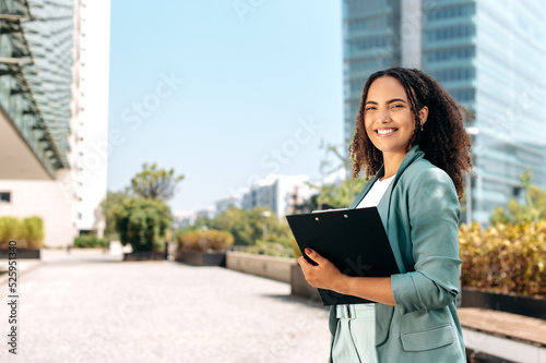 Successful positive confident young brazilian or hispanic curly haired business woman, real estate agent, manager, holds documents, stand outdoors near business center, looks at camera, smile friendly